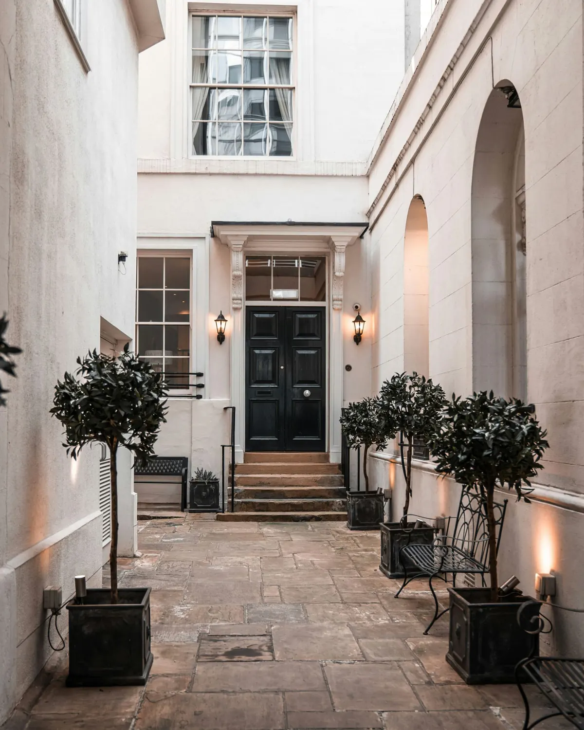 A flagged courtyard leading to a black panelled door with a fanlight, with sash windows above and bay trees in planters